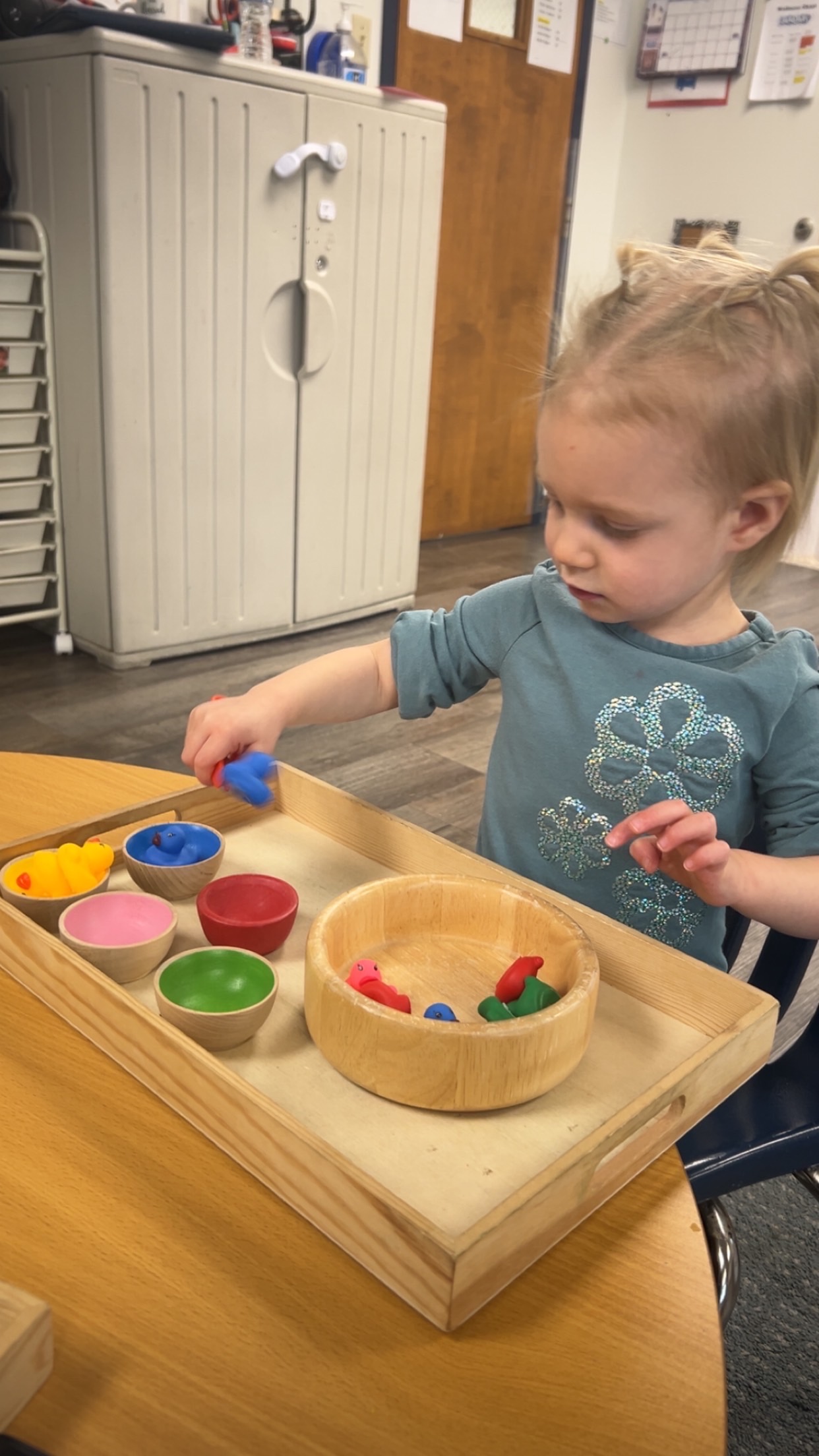 A toddler placing small rubber ducks into matching colored wooden bowls on a tray.